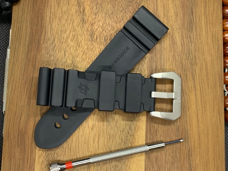 May include: Black rubber watch strap with a silver buckle and a small screwdriver. The strap is marked "SWISS MADE". The watch strap is lying on a wooden surface.