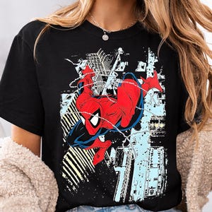 Puede incluir: Camiseta negra con un gr&aacute;fico de Spider-Man en una pose din&aacute;mica, con un traje rojo y azul, sobre un fondo de paisaje urbano. El dise&ntilde;o incluye detalles de telara&ntilde;a y un estilo de c&oacute;mic.
