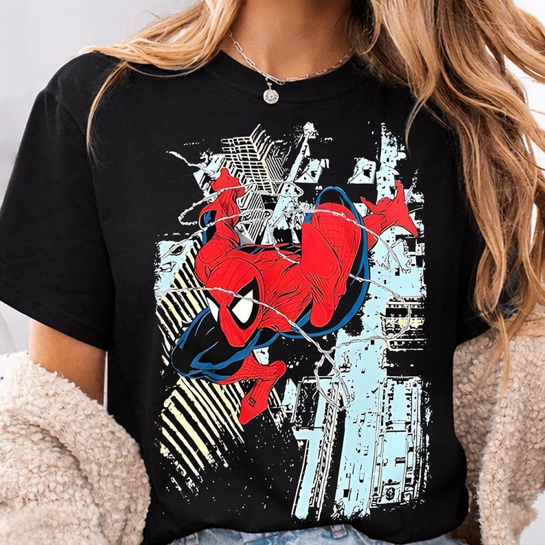 Puede incluir: Camiseta negra con un gr&aacute;fico de Spider-Man en una pose din&aacute;mica, con un traje rojo y azul, sobre un fondo de paisaje urbano. El dise&ntilde;o incluye detalles de telara&ntilde;a y un estilo de c&oacute;mic.