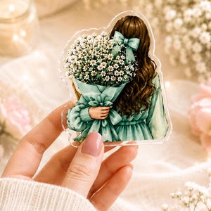May include: An acrylic charm featuring an illustration of a person with long brown hair tied with a light green bow, wearing a matching green dress and holding a large bouquet of white baby's breath flowers wrapped in green paper.