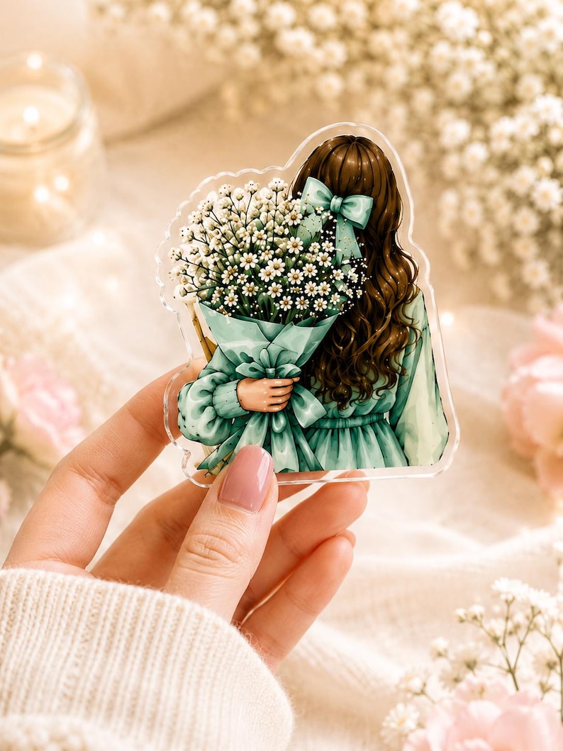 May include: An acrylic charm featuring an illustration of a person with long brown hair tied with a light green bow, wearing a matching green dress and holding a large bouquet of white baby's breath flowers wrapped in green paper.