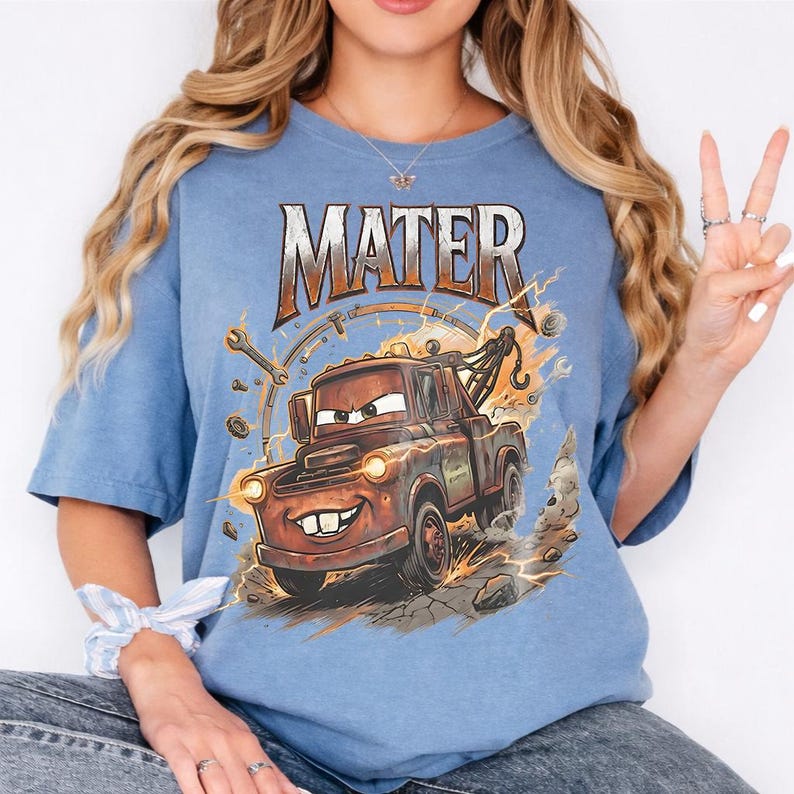 May include: A light blue t-shirt featuring the word "MATER" in a distressed, orange font above a cartoon image of Mater the tow truck. The graphic includes lightning bolts and tools, creating a dynamic design.