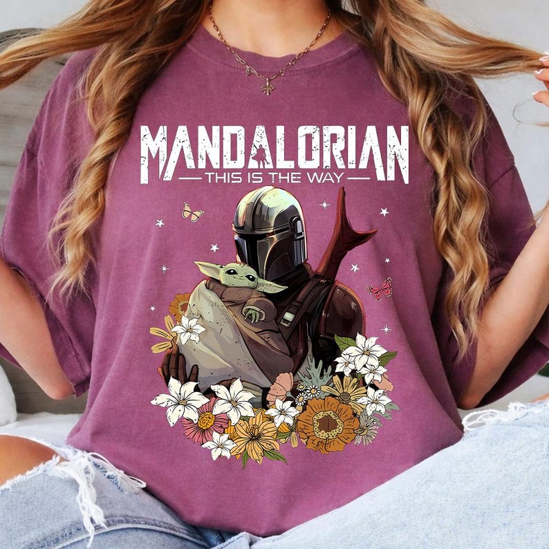 May include: A maroon t-shirt featuring a graphic of The Mandalorian and Grogu surrounded by flowers and butterflies. The text "MANDALORIAN - THIS IS THE WAY" is printed across the top in white.
