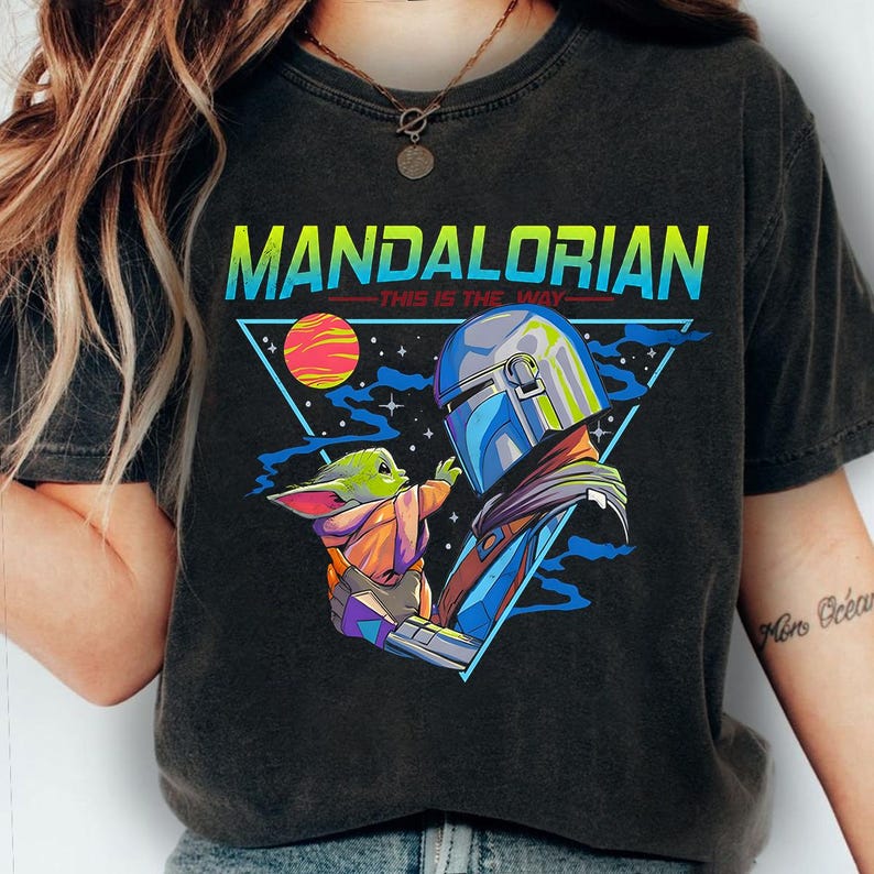 May include: Dark gray t-shirt featuring a colorful graphic of the Mandalorian and Grogu, with the text "MANDALORIAN - THIS IS THE WAY." The design includes a space scene with a planet and stars, and a triangular frame.