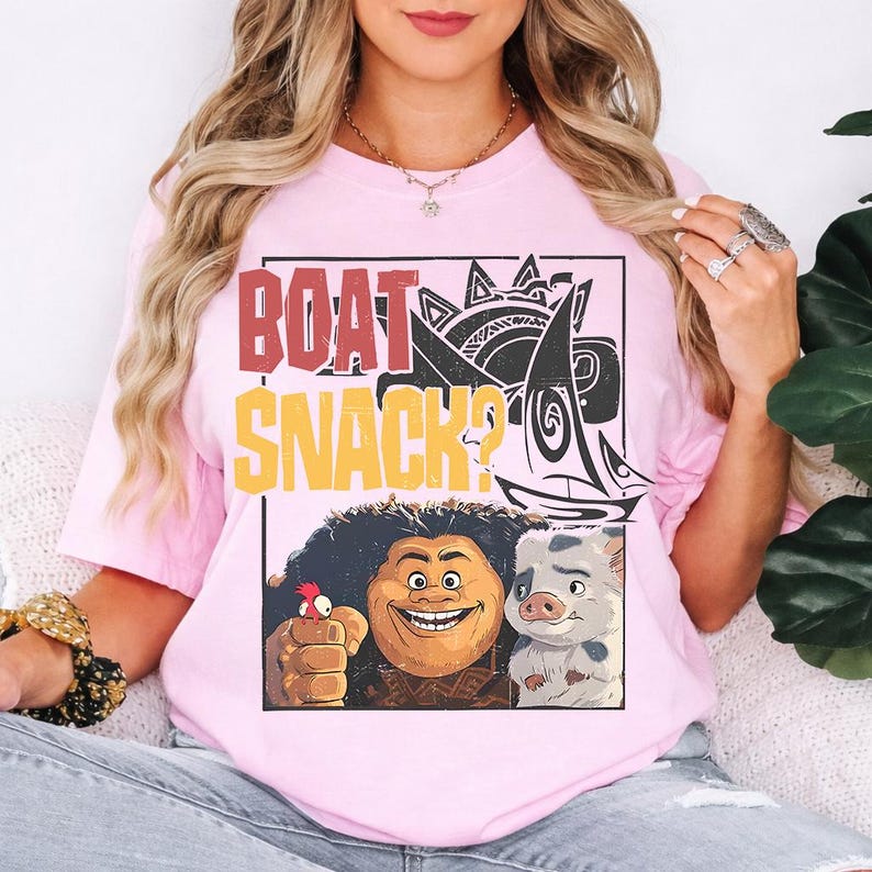 May include: A light pink short-sleeve t-shirt featuring a graphic print with the text BOAT SNACK? in red and yellow. The design includes a stylized sailboat, a cartoon character with dark curly hair, a small red bird, and a spotted piglet.