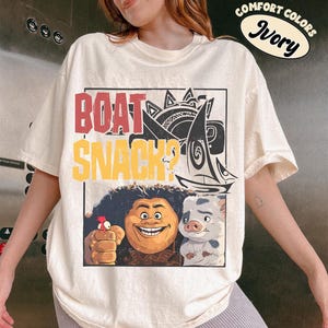 May include: An ivory short-sleeve t-shirt featuring a graphic print with the text "BOAT SNACK?" in red and yellow. The design includes a stylized sailboat, a smiling character with dark curly hair, a small red bird, and a spotted piglet character.