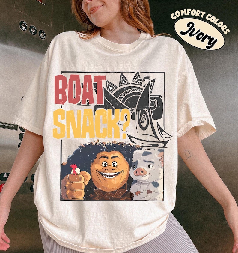 May include: An ivory short-sleeve t-shirt featuring a graphic print with the text "BOAT SNACK?" in red and yellow. The design includes a stylized sailboat, a smiling character with dark curly hair, a small red bird, and a spotted piglet character.