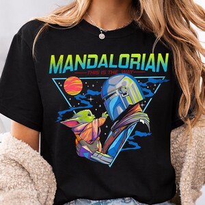 May include: Black t-shirt featuring a graphic of The Mandalorian and Grogu, with the text "MANDALORIAN - THIS IS THE WAY" in a triangle design. The illustration includes a planet, stars, and a space theme.