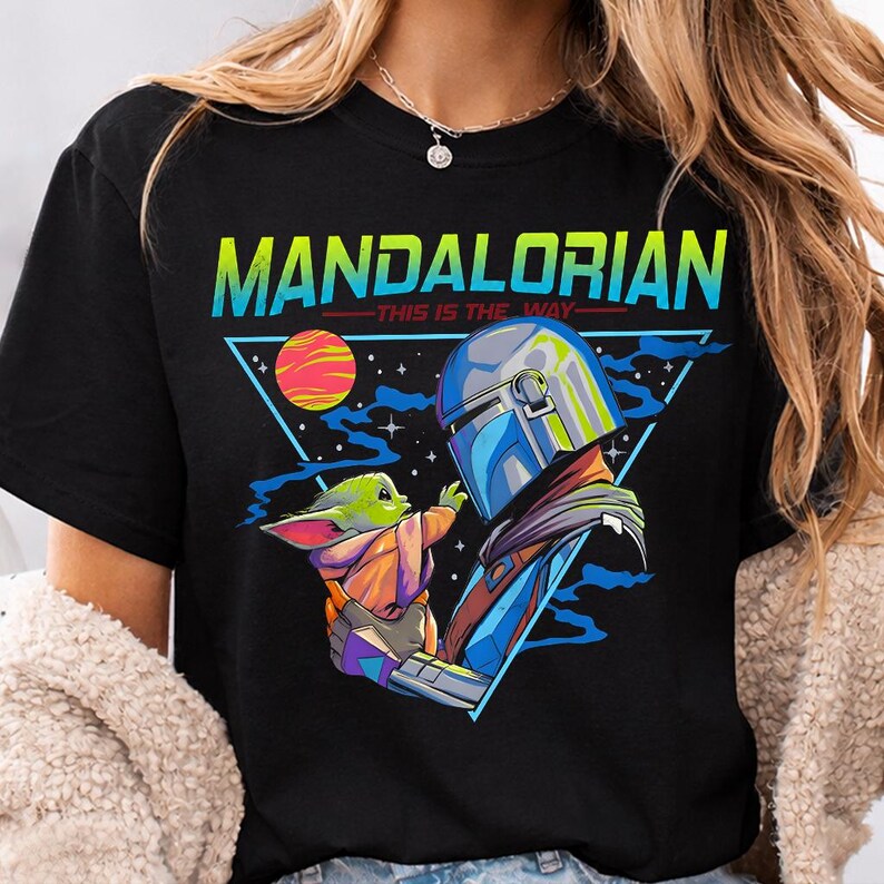 May include: Black t-shirt featuring a graphic of The Mandalorian and Grogu, with the text "MANDALORIAN - THIS IS THE WAY" in a triangle design. The illustration includes a planet, stars, and a space theme.