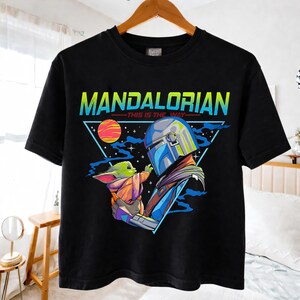 May include: Black t-shirt featuring a colorful graphic of The Mandalorian and Grogu. The design includes the text "MANDALORIAN" in yellow and blue, with "THIS IS THE WAY" below. The artwork depicts the characters in a triangular frame.