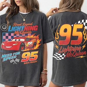 May include: Dark gray t-shirt featuring a graphic of Lightning McQueen from the movie Cars. The front design includes the text "Street Legend Lightning McQueen" and "Need for Speed 95 Race Ready". The back displays "95 Lightning McQueen".