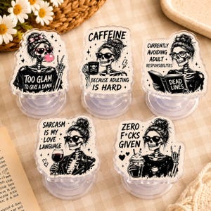 May include: Five clear phone grips featuring black and white illustrations of skeletons with messy buns and sunglasses. Each grip displays unique text: Too Glam To Give A Damn, Caffeine Because Adulting Is Hard, Currently Avoiding Adult Responsibilities, Sarcasm Is My Love Language, and Zero F*cks Given.