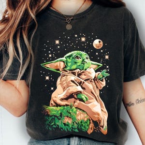 May include: Dark gray t-shirt featuring a green cartoon character with large ears, wearing a tan robe, and set against a starry background with a planet. The character is reaching out with one hand.