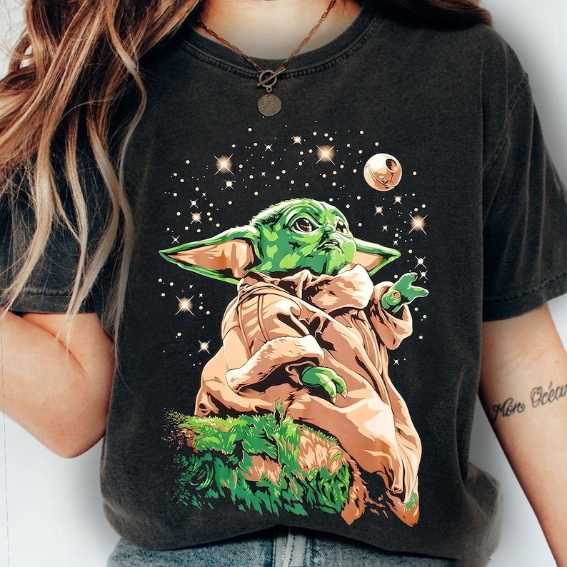 May include: Dark gray t-shirt featuring a green cartoon character with large ears, wearing a tan robe, and set against a starry background with a planet. The character is reaching out with one hand.