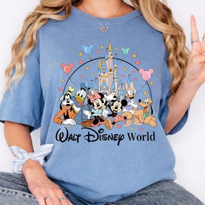 K&ouml;nnte beinhalten: Hellblaues T-Shirt mit einer Grafik von Disney-Figuren, darunter Micky und Minnie Maus, Donald Duck und Goofy, vor einem Schloss. Die Worte "Walt Disney World" sind unter den Figuren aufgedruckt.