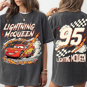 May include: Dark gray t-shirt featuring Lightning McQueen from the Cars movie. The front graphic shows McQueen racing with lightning bolts and the text "Lightning McQueen". The back displays the number "95" with a checkered flag.