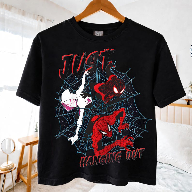 Puede incluir: Camiseta negra con un dise&ntilde;o de Spider-Man. El dise&ntilde;o presenta a Spider-Man, Spider-Gwen y otra figura de Spider-Man, todo sobre un fondo de telara&ntilde;a. Las palabras "JUST" y "HANGING OUT" son rojas.