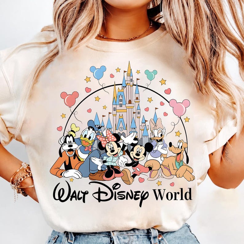 K&ouml;nnte beinhalten: Cremefarbenes Sweatshirt mit einer Grafik von Disney-Figuren, darunter Mickey und Minnie Mouse, Donald Duck und Goofy, vor einem Schloss. Die Worte "Walt Disney World" sind unter den Figuren in einer Schriftart aufgedruckt. Ballons und Sterne sind ebenfalls enthalten.