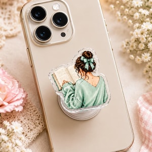 May include: A light-colored smartphone with a circular phone grip attached to the back. The grip features an illustration of a person with a brown hair bun and a teal bow, wearing a mint green sweater while reading an open book.