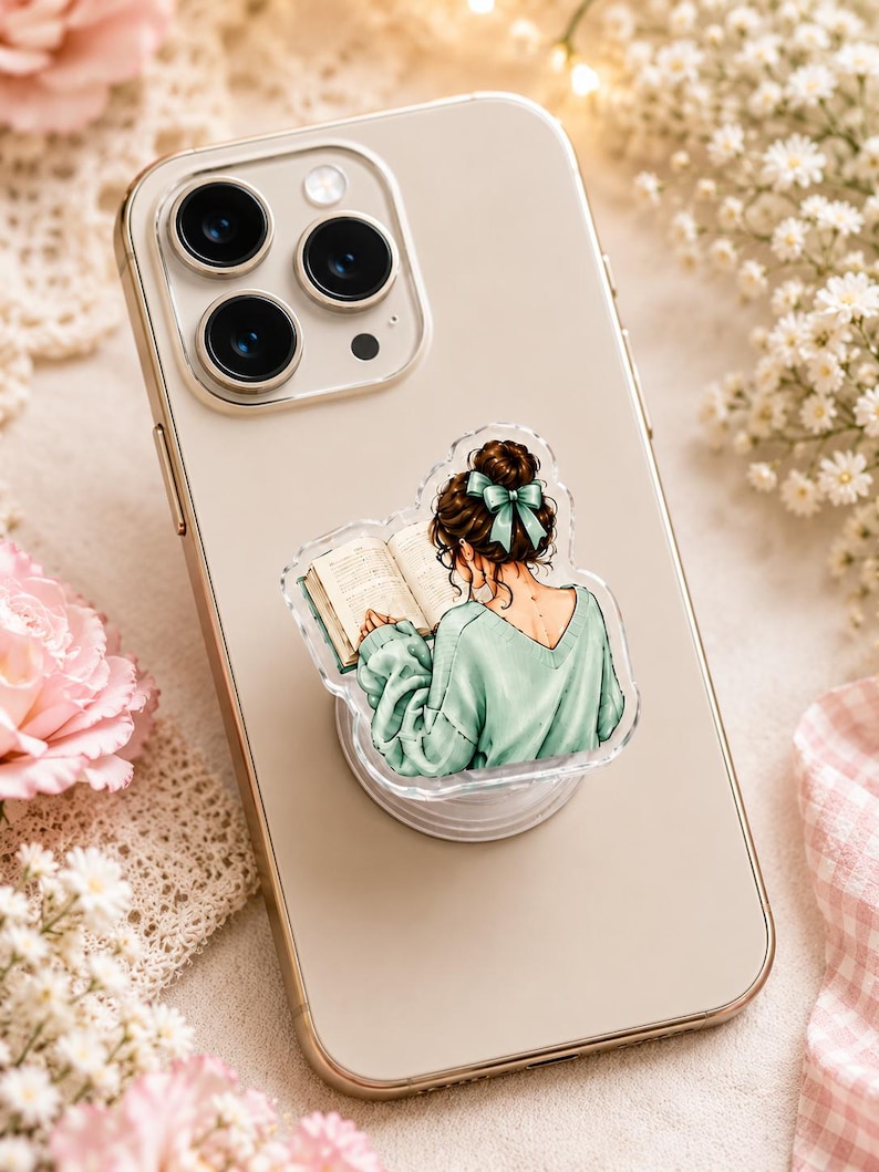 May include: A light-colored smartphone with a circular phone grip attached to the back. The grip features an illustration of a person with a brown hair bun and a teal bow, wearing a mint green sweater while reading an open book.