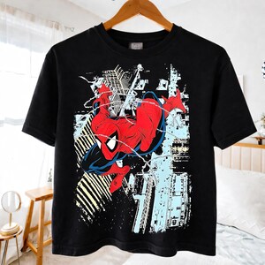 Puede incluir: Camiseta negra con un gr&aacute;fico de Spider-Man en una pose din&aacute;mica, con detalles en rojo y azul. El dise&ntilde;o incluye elementos de la ciudad y telara&ntilde;as. La camiseta est&aacute; colgada en una percha de madera.