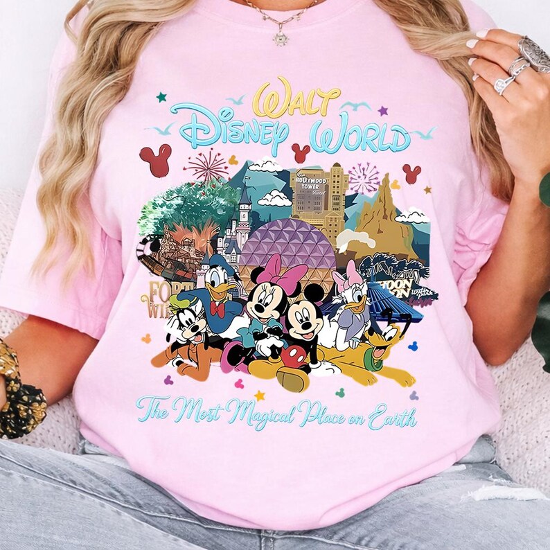 K&ouml;nnte beinhalten: Hellrosa T-Shirt mit einem farbenfrohen Aufdruck von Disney-Figuren und Wahrzeichen, darunter Micky und Minnie Maus, Donald Duck und das Hollywood Tower Hotel. Der Text lautet "Walt Disney World" und "The Most Magical Place on Earth."