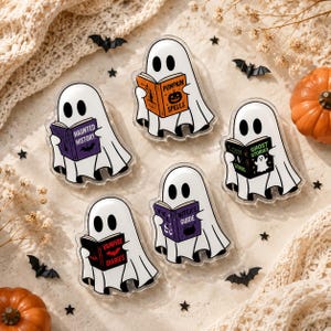 May include: Five acrylic pins featuring white ghosts reading books. The books are titled Haunted History, Pumpkin Spells, Ghost Stories, Vampire Diaries, and Witch's Guide. Each ghost has black oval eyes and is depicted in a cartoon style.