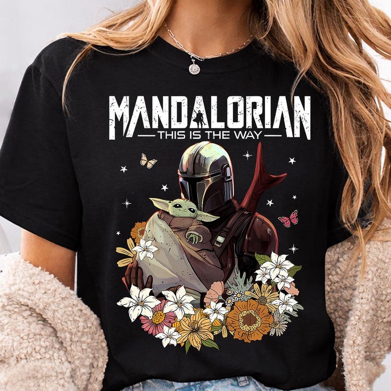 May include: Black t-shirt featuring a graphic of The Mandalorian holding Grogu, surrounded by flowers and butterflies. The text "MANDALORIAN - THIS IS THE WAY" is printed across the top in white.