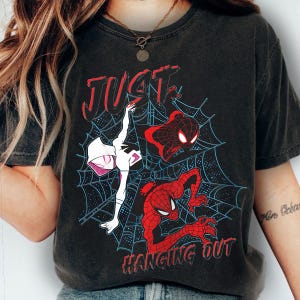 Puede incluir: Camiseta negra con un dise&ntilde;o de personajes de Spider-Man en una telara&ntilde;a. El texto "JUST HANGING OUT" es rojo. El dise&ntilde;o incluye a Spider-Gwen y dos figuras de Spider-Man.