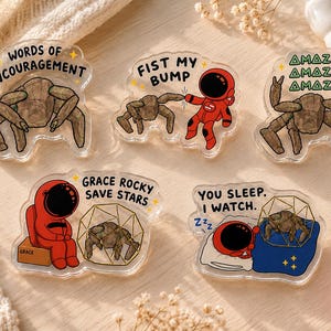May include: Five clear acrylic pins featuring cartoon illustrations of a brown rock-like creature and a red astronaut. The text includes: WORDS OF ENCOURAGEMENT, FIST MY BUMP, AMAZE AMAZE AMAZE, GRACE ROCKY SAVE STARS, and YOU SLEEP. I WATCH. with small yellow stars.