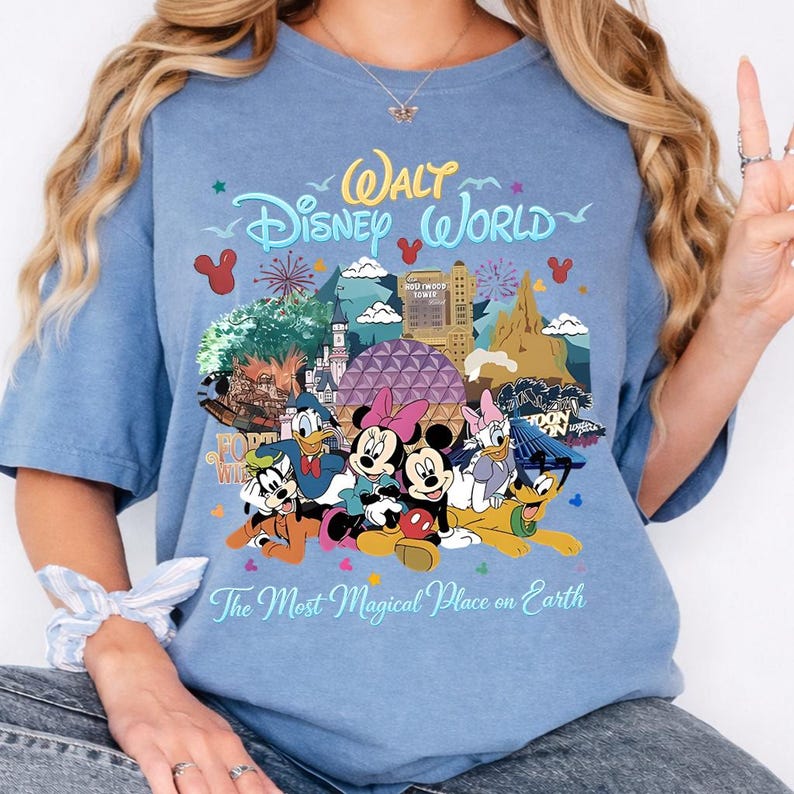 K&ouml;nnte beinhalten: Hellblaues T-Shirt mit einem farbenfrohen Grafikdesign von Walt Disney World-Charakteren und -Sehensw&uuml;rdigkeiten. Das Design zeigt Micky und Minnie Maus, Donald Duck und Goofy, mit dem Text "Walt Disney World" und "The Most Magical Place on Earth."