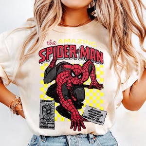 May include: A cream-coloured t-shirt with a comic book graphic of Spider-Man. The design shows Spider-Man in a crouching pose, with the text "the AMAZING SPIDER-MAN" in red and yellow, and other comic book elements.