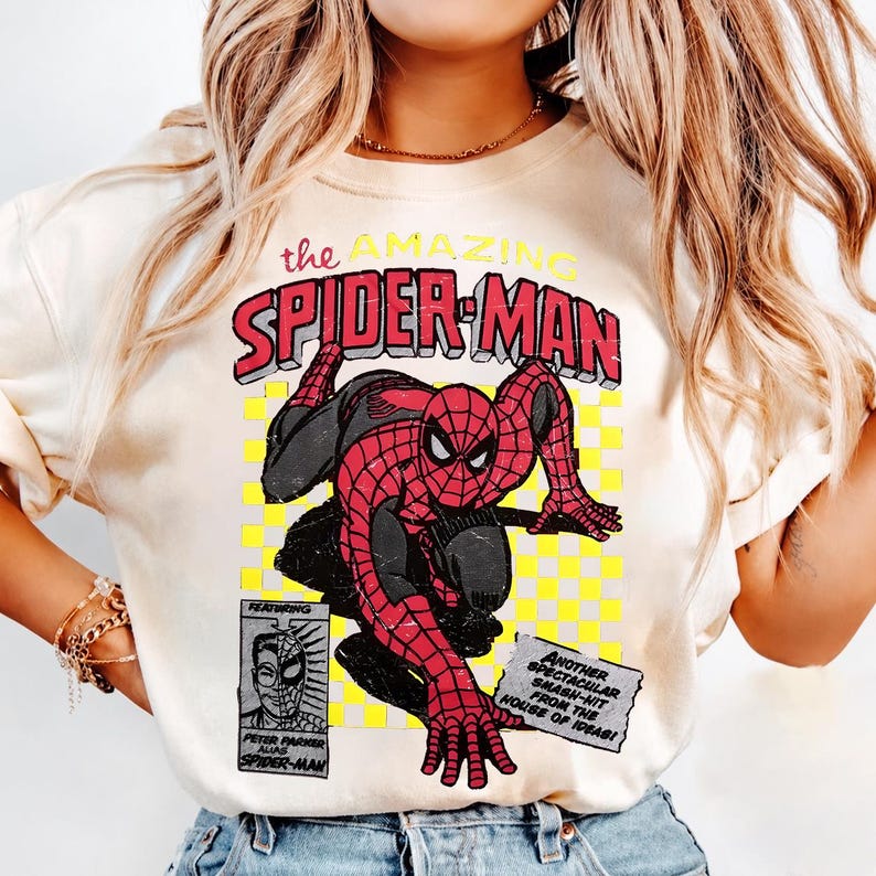 May include: A cream-coloured t-shirt with a comic book graphic of Spider-Man. The design shows Spider-Man in a crouching pose, with the text "the AMAZING SPIDER-MAN" in red and yellow, and other comic book elements.