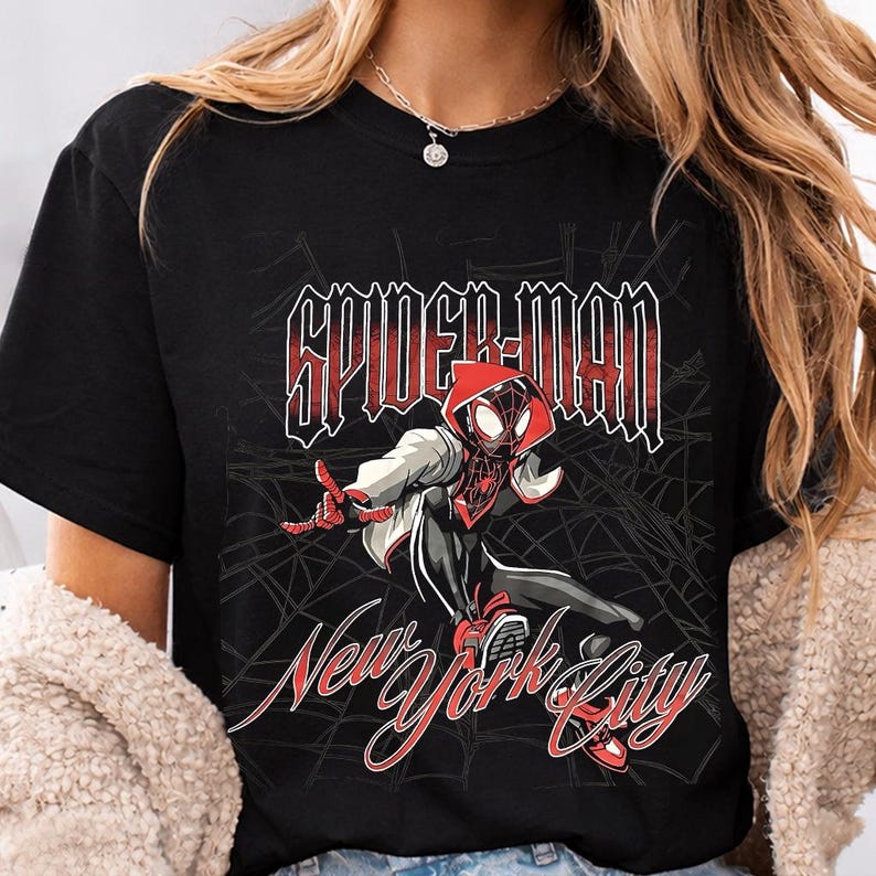 May include: Black t-shirt featuring a graphic of Spider-Man in a dynamic pose. The design includes the text "SPIDER-MAN" in a bold, outlined font above "New York City" in a cursive script, all in red. A spider web design is in the background.