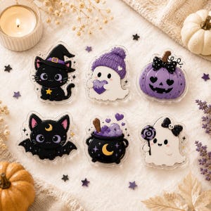 May include: A set of six acrylic pins featuring Halloween-themed illustrations: a black cat in a witch hat, a white ghost in a purple beanie holding a heart, a purple jack-o'-lantern, a black bat-cat, a black cauldron, and a white ghost with a lollipop.
