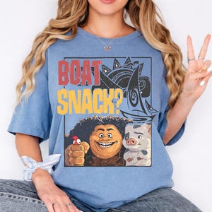 May include: A blue short-sleeve t-shirt featuring a graphic print with the text "BOAT SNACK?" in red and yellow. The design includes a stylized sailboat, a cartoon character with dark curly hair, a small red bird, and a piglet character in a frame.
