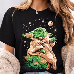 May include: Black t-shirt featuring a green alien character with large ears, wearing a tan robe, set against a starry night sky with a moon. The character is seated on green foliage.