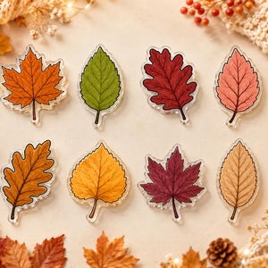 Op de afbeelding: Een set van acht bladvormige acryl ornamenten met diverse botanische ontwerpen. De collectie bevat vormen van esdoorn-, eiken- en berkenbladeren in levendige herfstkleuren zoals oranje, groen, donkerrood, roze, bruin, geel en donkerpaars.