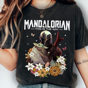 May include: Dark gray t-shirt featuring the text "MANDALORIAN - THIS IS THE WAY" above an illustration of the Mandalorian holding Grogu surrounded by flowers and butterflies. The design includes a helmet, Grogu, and floral elements.