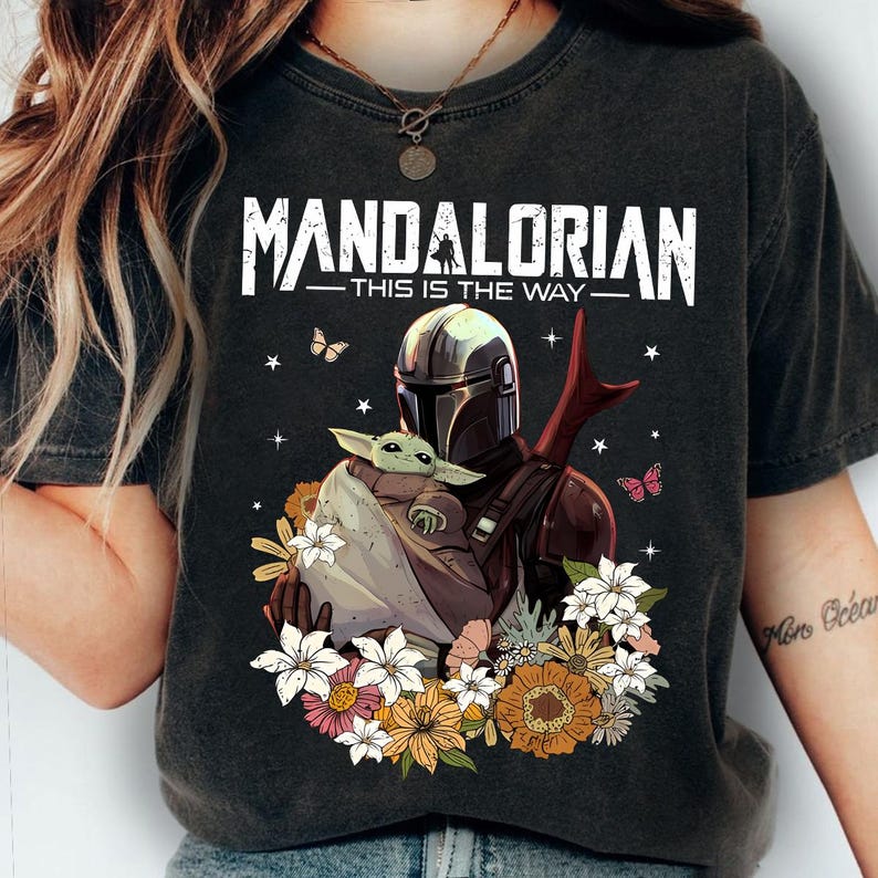 May include: Dark gray t-shirt featuring the text "MANDALORIAN - THIS IS THE WAY" above an illustration of the Mandalorian holding Grogu surrounded by flowers and butterflies. The design includes a helmet, Grogu, and floral elements.
