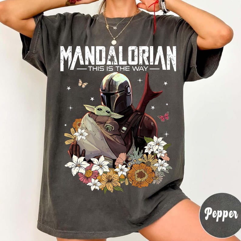 May include: Dark gray t-shirt featuring a graphic of The Mandalorian holding Grogu, surrounded by flowers and butterflies. The text "MANDALORIAN - THIS IS THE WAY" is printed across the top in white.