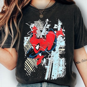 Puede incluir: Camiseta gris oscuro con un gr&aacute;fico de Spider-Man en una pose din&aacute;mica. El superh&eacute;roe est&aacute; representado en su cl&aacute;sico traje rojo y azul, con telara&ntilde;as y un fondo de paisaje urbano en blanco, azul claro y amarillo.