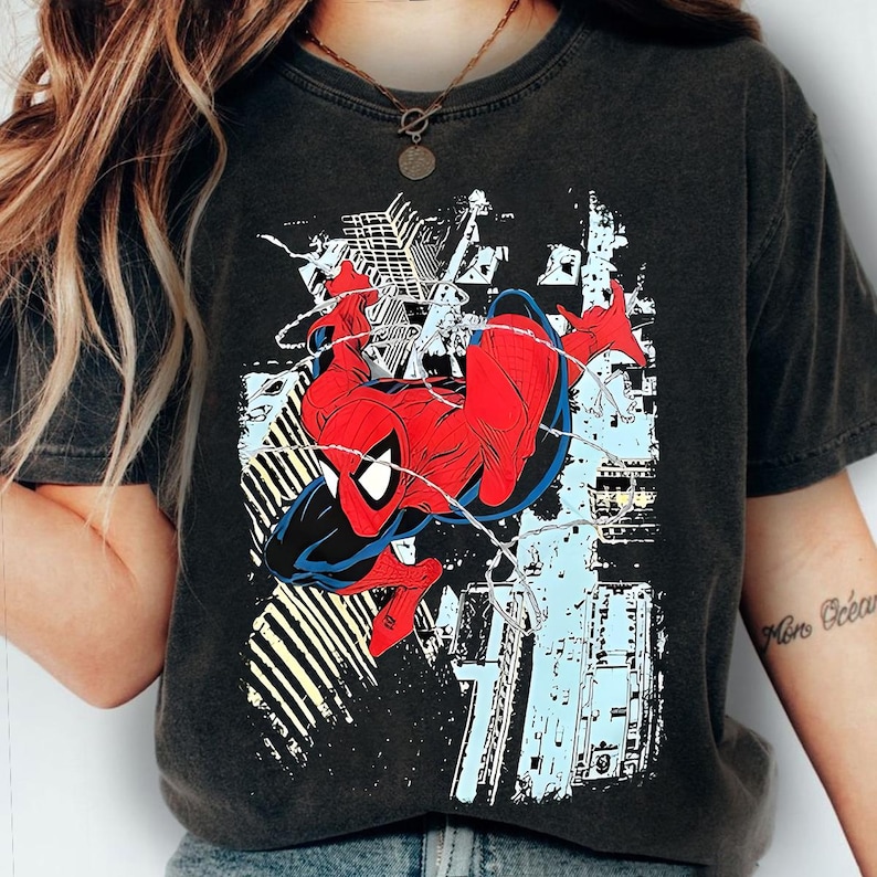 Puede incluir: Camiseta gris oscuro con un gr&aacute;fico de Spider-Man en una pose din&aacute;mica. El superh&eacute;roe est&aacute; representado en su cl&aacute;sico traje rojo y azul, con telara&ntilde;as y un fondo de paisaje urbano en blanco, azul claro y amarillo.