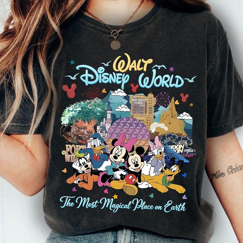 K&ouml;nnte beinhalten: Dunkelgraues T-Shirt mit einer farbenfrohen Grafik von Disney-Figuren und -Sehensw&uuml;rdigkeiten, darunter Micky und Minnie Maus, Donald Duck und das Hollywood Tower Hotel. Der Text "Walt Disney World" und "The Most Magical Place on Earth" ist ebenfalls abgebildet.