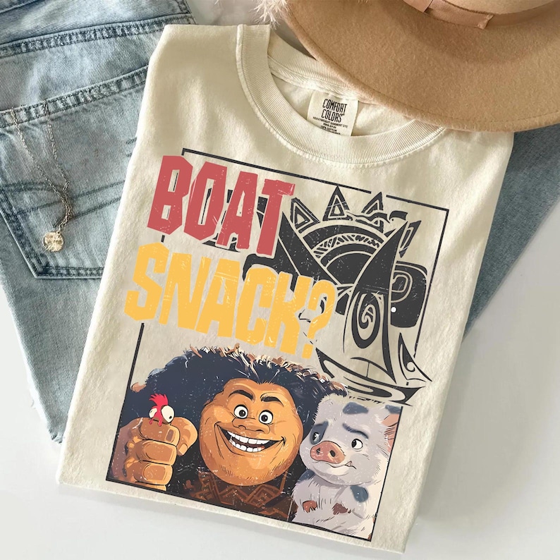 May include: A cream-colored t-shirt featuring a graphic print with the text "BOAT SNACK?" in red and yellow. The design includes illustrations of a man with curly hair, a small chicken, a pig, and a sailboat within a rectangular black frame.