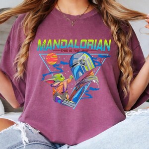 May include: A maroon t-shirt featuring a graphic design with the text "MANDALORIAN - THIS IS THE WAY". The design includes an illustration of the Mandalorian and Grogu, with a colorful sun and triangle background.