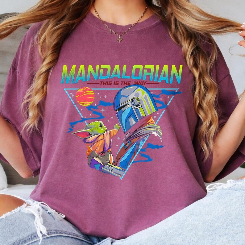May include: A maroon t-shirt featuring a graphic design with the text "MANDALORIAN - THIS IS THE WAY". The design includes an illustration of the Mandalorian and Grogu, with a colorful sun and triangle background.