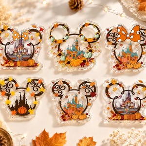 Op de afbeelding: Zes doorzichtige acryl ornamenten in de vorm van muizenoortjes, elk met een gedetailleerde kasteelillustratie omringd door herfstbladeren, pompoenen en zonnebloemen. Sommige ontwerpen bevatten oranje strikken met stippen.