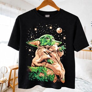 May include: Black t-shirt featuring a graphic of a green alien character with large ears, wearing a tan robe, and set against a starry night sky with a moon. The character is sitting on a green, mossy surface.