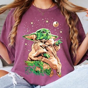 May include: A maroon t-shirt featuring a graphic of a green alien character with large ears, wearing a tan robe, and set against a starry background with a moon. The character is positioned on a patch of green foliage.